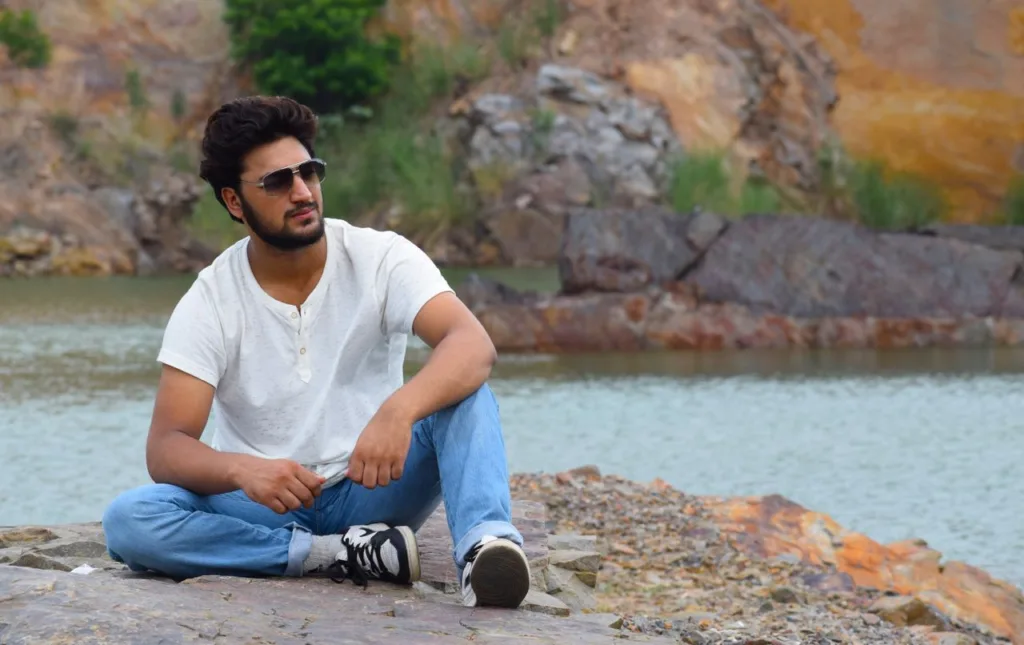 Casual young man in sunglasses sitting on rocks by a serene lakeside, showcasing relaxed outdoor lifestyle.