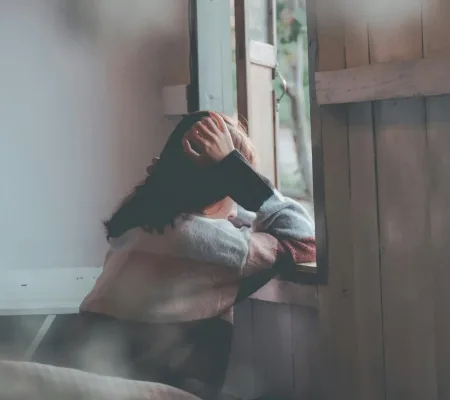 A woman sits indoors gazing thoughtfully out of a window, evoking emotions of loneliness and reflection.