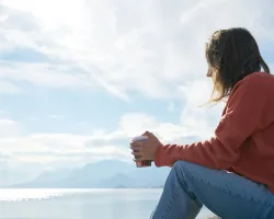 A woman in casual attire enjoys a hot drink while gazing at the sea, epitomizing relaxation.