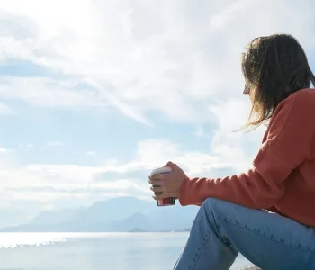 A woman in casual attire enjoys a hot drink while gazing at the sea, epitomizing relaxation.