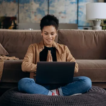 A smiling young woman sits cross-legged on a pouf, using her laptop in a cozy home setting.